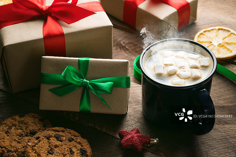 准备一个家庭节日的概念，平安夜快乐。用鲜红的丝带捆扎好圣诞礼物，盒子上挂着蝴蝶结，背景是一张木桌。杯子里有热咖啡和棉花糖，饼干和巧克力。图片素材