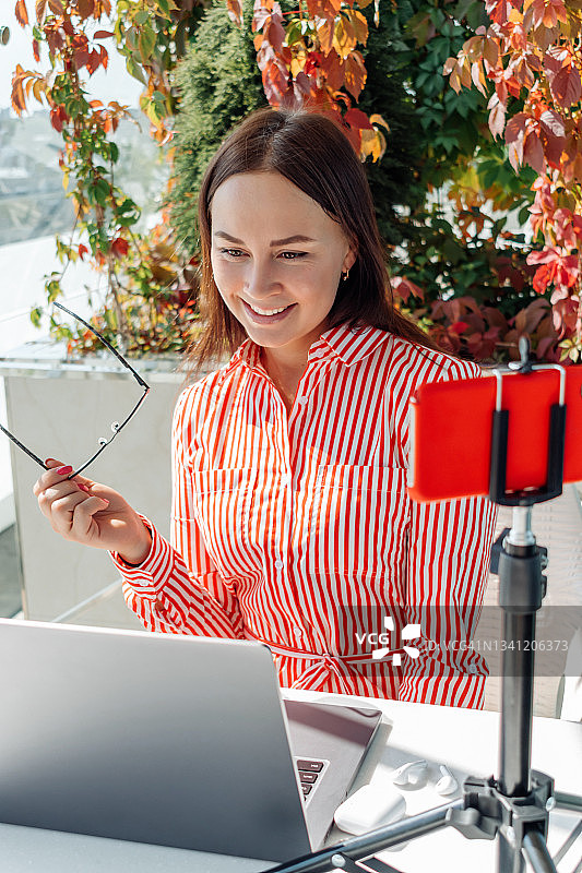 一个夏天，年轻的黑发女商人戴着眼镜坐在咖啡馆的笔记本电脑屏幕前，通过智能手机进行视频电话会议。业务,电子学习的概念。图片素材