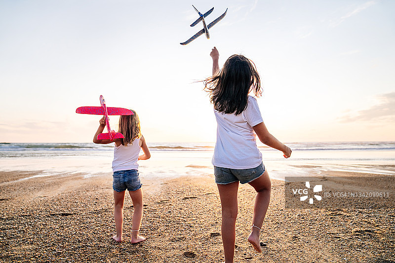 幸福童年的概念。两姐妹在夕阳下跑着玩着玩具飞机。孩子们在阳光明媚的背景下，手里拿着飞机。孩子们把玩具飞机扔向天空的剪影图片素材