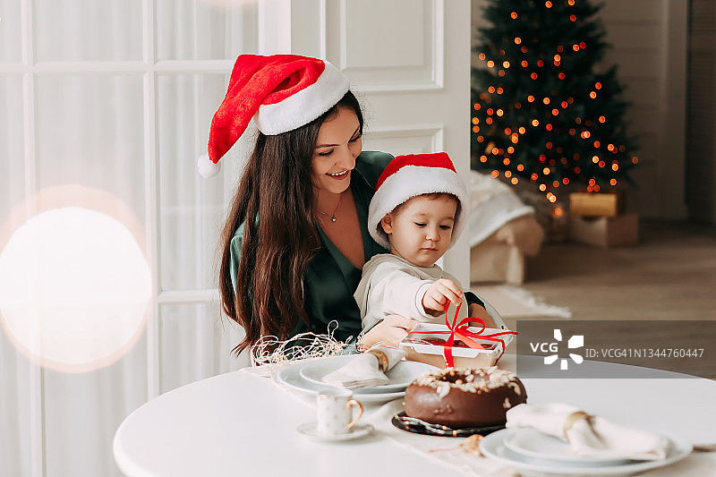 圣诞节的概念。快乐活泼的年轻妈妈和戴着红色圣诞老人帽子的小儿子坐在节日餐桌前庆祝寒假，妈妈祝贺送给孩子一个礼品盒，房间里装饰着一棵圣诞树图片素材