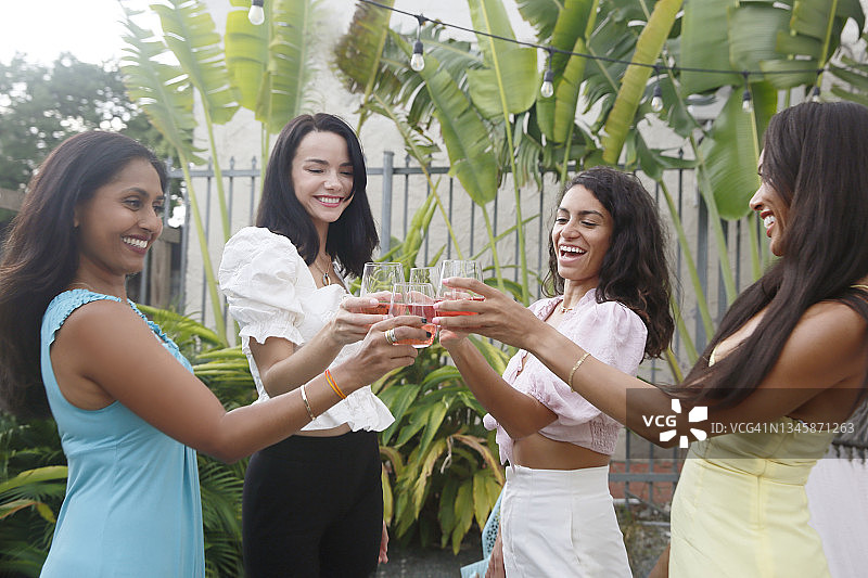 一个混合种族的妇女团体，在室外庭院的一个小型庆祝活动中享受葡萄酒祝酒词，穿着休闲的聚会服装。图片素材