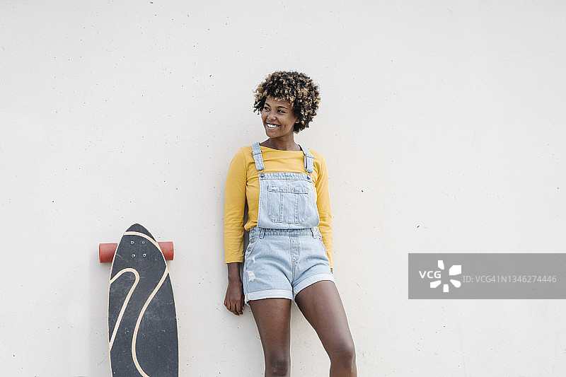 穿着都市风格服装的非洲式女滑冰选手，倚在户外的墙上微笑。图片素材