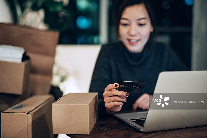年轻的亚洲女性用笔记本电脑在网上购物，在家用信用卡进行移动支付。她身边还有购物袋和送货箱。科技让生活变得简单多了图片素材