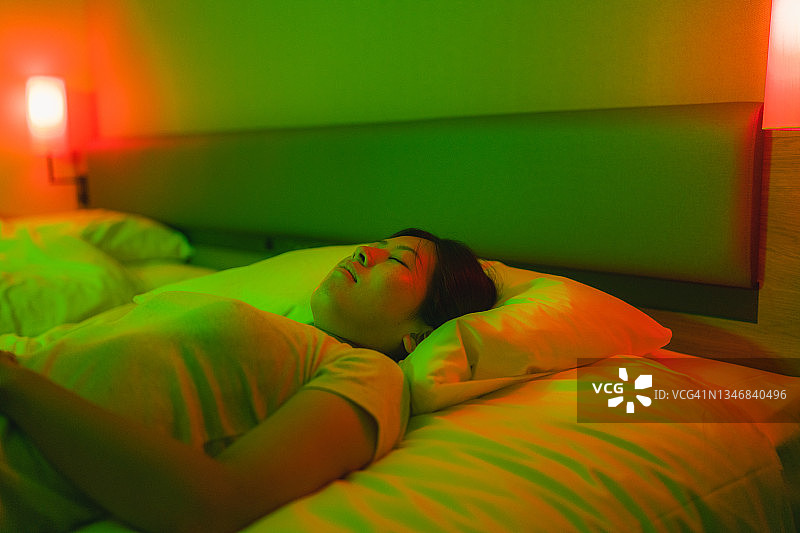 年轻女子被红色和绿色的霓虹灯照亮，躺在家里卧室的床上休息图片素材