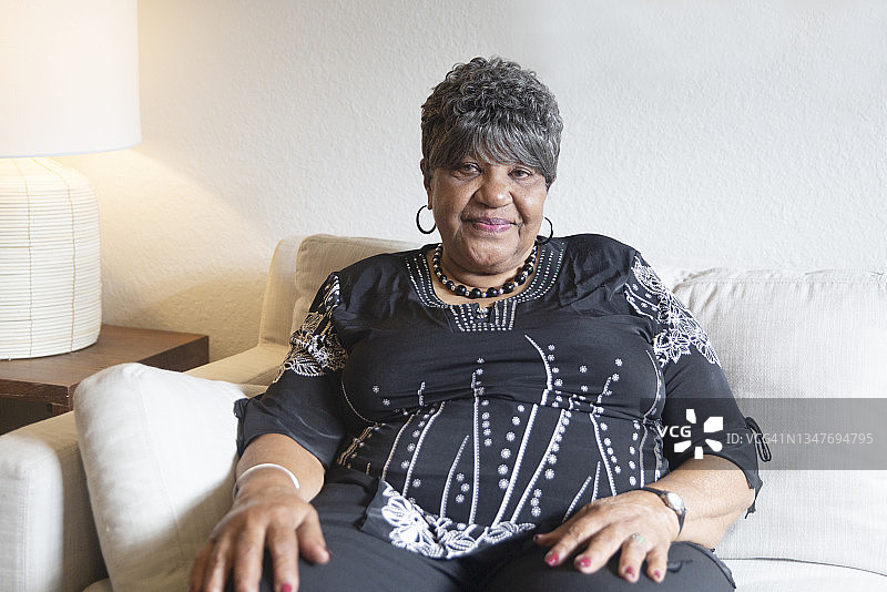 在明亮的客厅里，一位灰白头发的非裔美国老年妇女身穿黑白衬衫坐在沙发上。图片素材