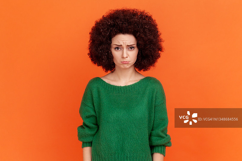 一个忧郁的女人，留着非洲式发型，穿着绿色休闲风格的毛衣，撅着嘴看着镜头，对坏消息感到沮丧。图片素材