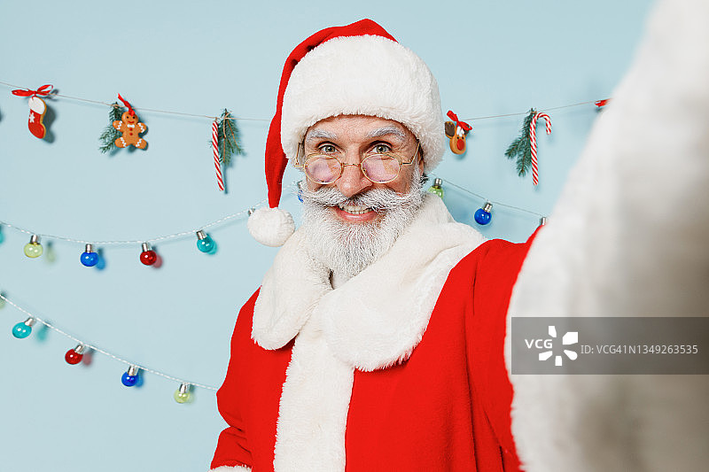 近距离观察满脸胡须的老人，50多岁，戴着圣诞帽，身穿红色套装，用手机自拍，在纯蓝色背景摄影棚，新年快乐，2022庆祝，圣诞快乐，圣诞概念。图片素材