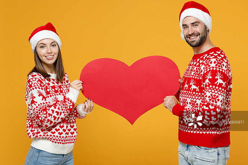 微笑着年轻的圣诞老人夫妇的朋友男人女人20多岁穿着红色的毛衣，圣诞帽hold大空空如也的心孤立在黄色的背景摄影棚。新年庆祝快乐节日概念。图片素材