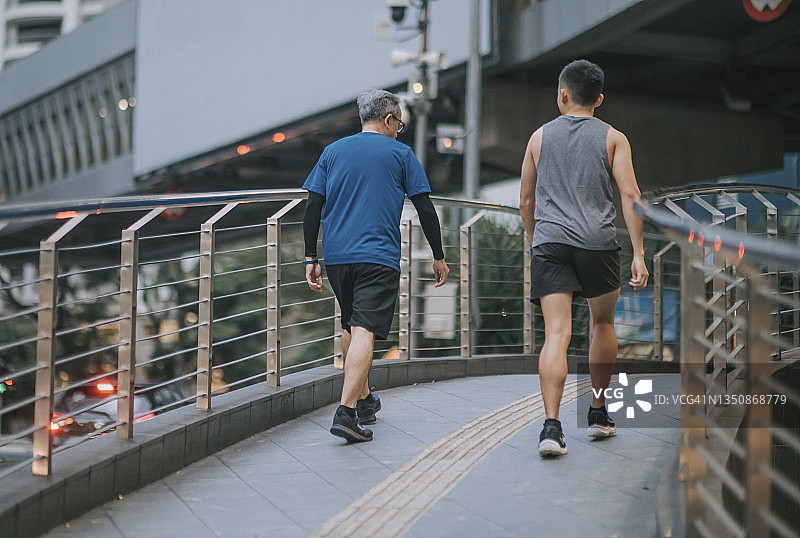 后视图亚洲华人活跃的老人和儿子晚上下班后一起在城里跑步慢跑图片素材