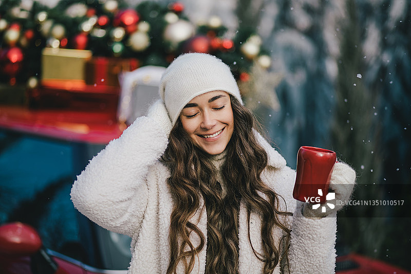 快乐美丽的年轻女子穿着针织毛衣和羊毛帽喝热巧克力附近的红色汽车与圣诞树在下雪。图片素材