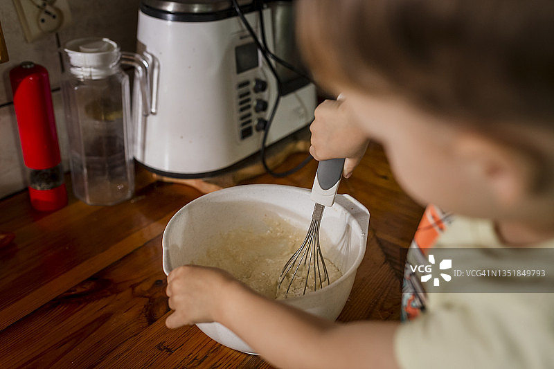 一个蹒跚学步的男孩正在白色的碗里搅拌奶油蛋糕图片素材
