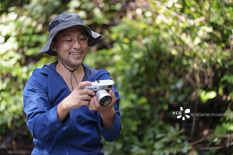 一名日本男子在热带森林里拿着相机看着照片，享受着假期。图片素材