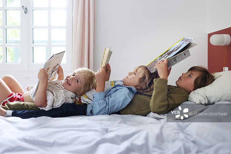 3个兄弟姐妹在家里的一张床上一起读书图片素材