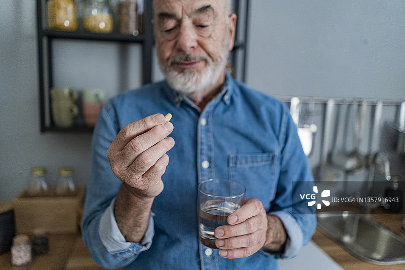 一位退休男子在厨房里拿着一杯水看着药丸图片素材