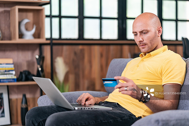 中年秃头男子坐在家里的沙发上，拿着笔记本电脑，用信用卡在网上购物和支付图片素材