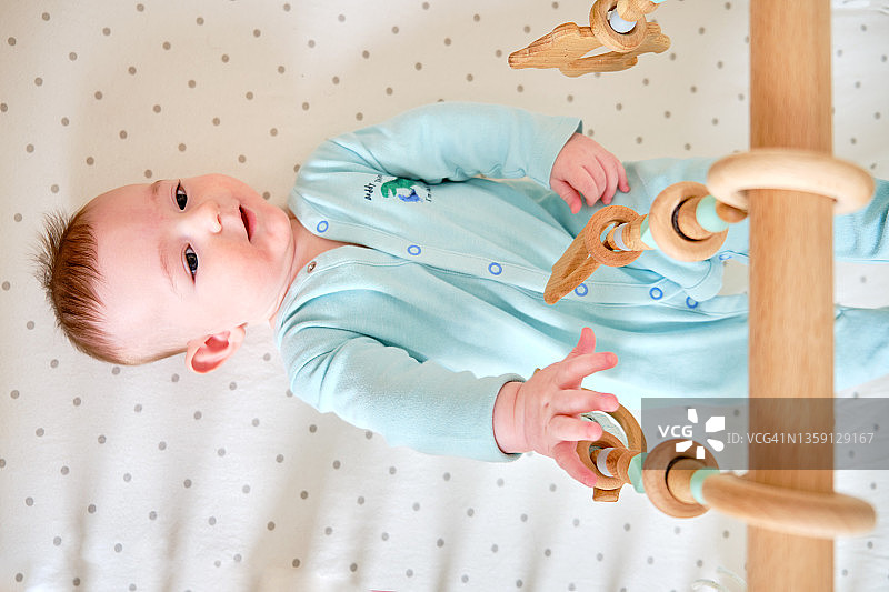 快乐的婴儿躺在床上玩着木制的吊玩具。6个月大的婴儿床里一个穿着绿松石衣服笑得滑稽的孩子图片素材