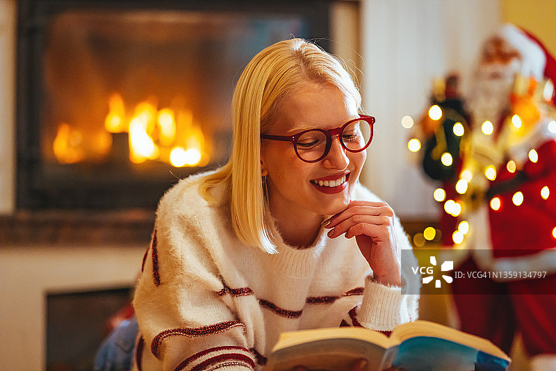 美丽的微笑金发女人拿着书，看着圣诞树。图片素材