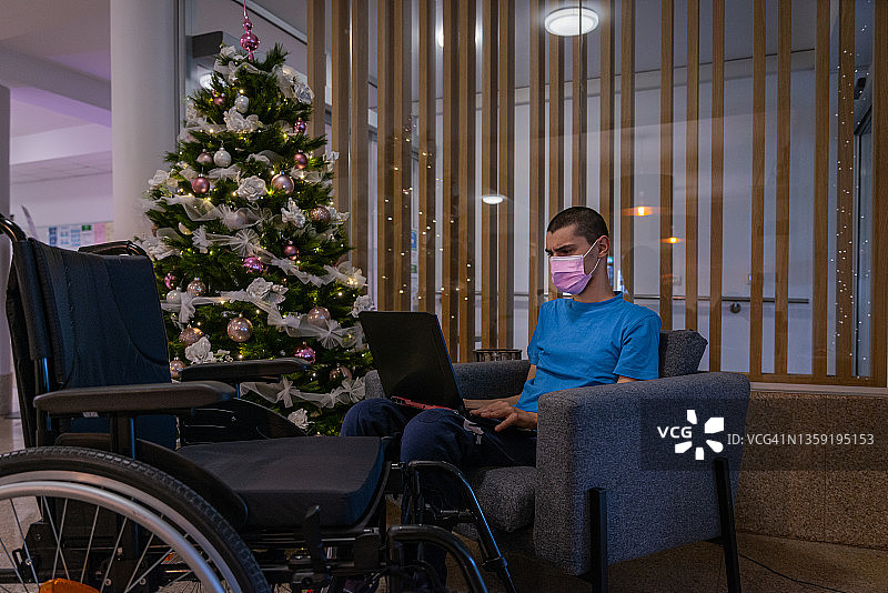 酒店大厅里，一棵装饰华丽、灯火通明的圣诞树旁，一名身患残疾、身穿蓝色t恤、因新冠肺炎而患有营养不良的青少年，带着医用口罩，坐在轮椅上。他一边工作，一边打电话，一边看着圣诞树图片素材