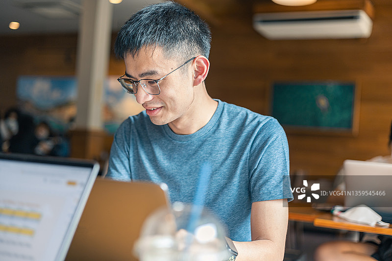 一个聪明的年轻人坐在繁忙的商务咖啡馆里，一边吃一边用笔记本电脑工作。图片素材