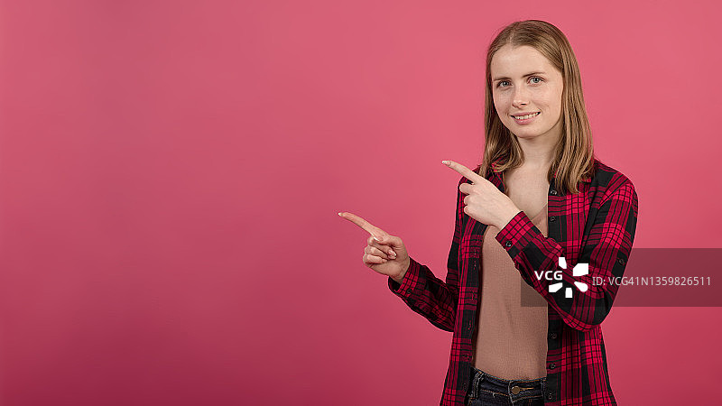 一个年轻迷人的金发白种人女孩，穿着衬衫，用手指指着粉色工作室背景上的一个空广告位。图片素材