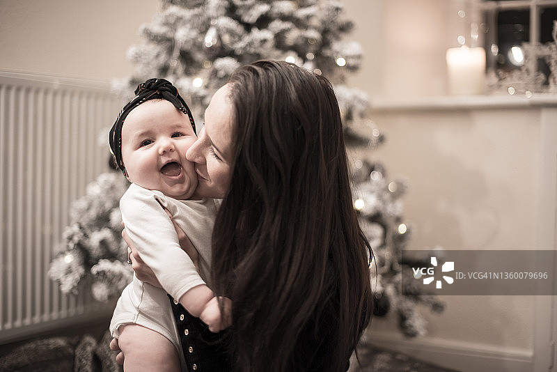 在英国苏格兰边境梅尔罗斯的一所房子里，一位年轻的母亲张开嘴，拥抱并亲吻她的孩子的脸颊，享受着圣诞树旁的拥抱图片素材