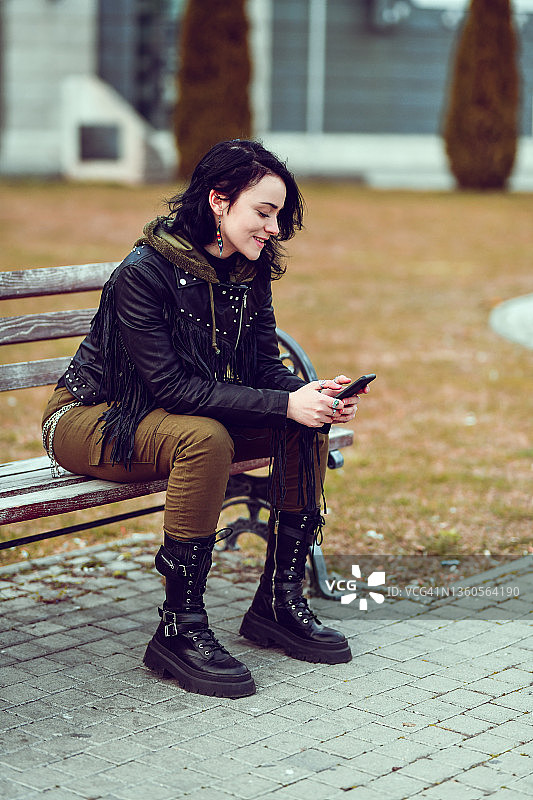 美丽的微笑哥特式女性使用智能手机，而放松在长凳外面图片素材