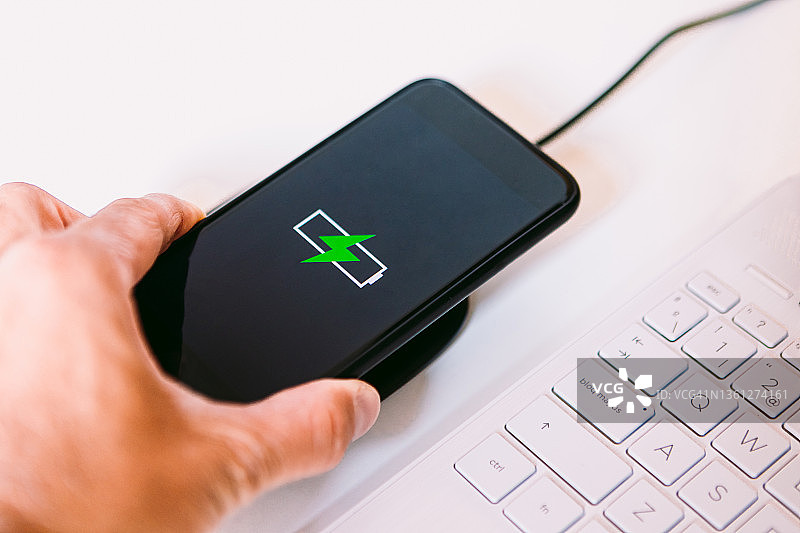 手持黑色智能手机，屏幕上有一个绿色闪电的电池标识，在白色工作台上笔记本电脑旁边的无线充电器底座上充电图片素材