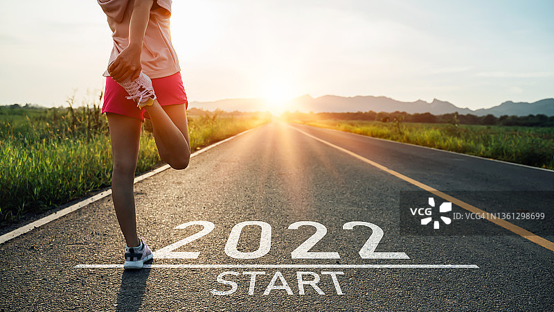 2022年新年或开始直观。沥青路上写着“2022”字样，日落时分，女运动员伸腿准备迎接新年。对挑战或职业道路和改变的概念。图片素材
