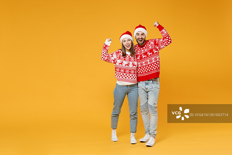 全长快乐年轻的圣诞老人夫妇朋友，男人女人在毛衣，圣诞帽做赢家姿态拥抱孤立在黄色背景工作室的肖像。祝新年欢度佳节理念。图片素材