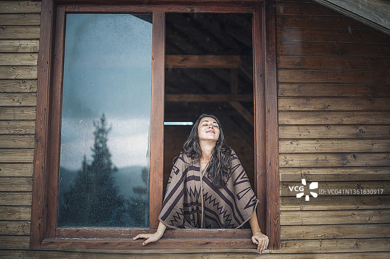 女人闭上眼睛享受大自然的清新，站在木屋的窗户上感受大自然的心情。图片素材