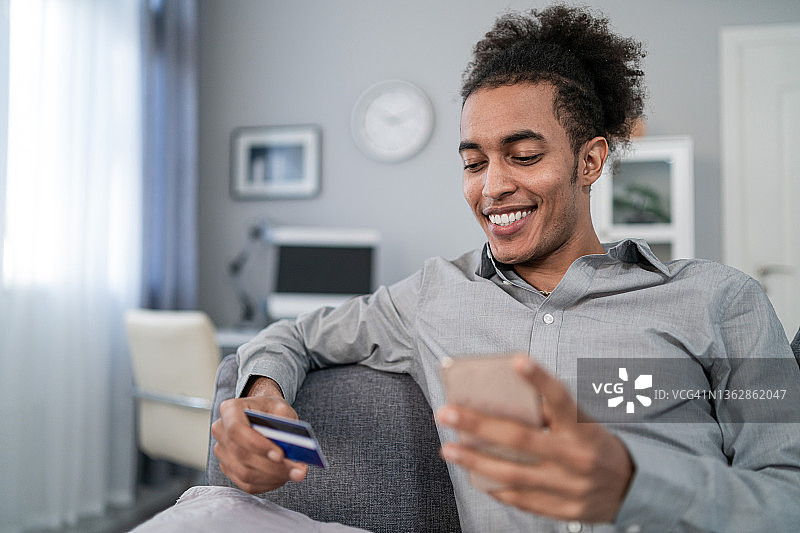 留着非洲式发型的多种族青年男子坐在客厅的沙发上，手里拿着手机和信用卡，通过手机app进行网上购物。图片素材