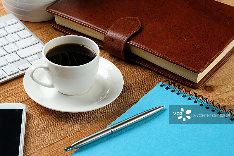 一个手机，一个电脑键盘，一支笔和一个笔记本，一个咖啡杯在一个木桌上。工作场所的商人或经理的主题。商务、咖啡和早餐的概念。图片素材
