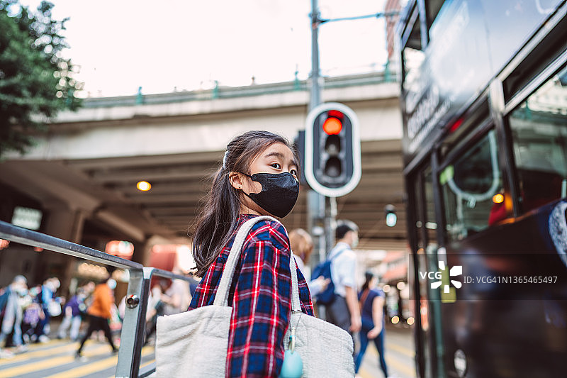 戴着防护口罩的可爱女孩穿过繁忙的城市街道，背景是公共汽车、交通和行人图片素材