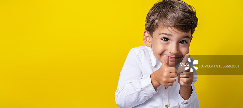 前视图是一个四岁的白人小男孩，站在黄色背景的工作室前微笑着，举着大拇指支持孩子和成功的概念图片素材