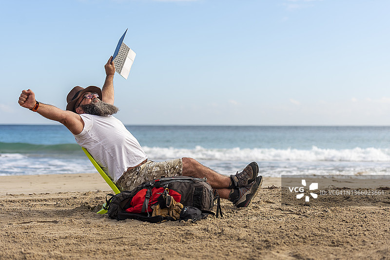 留着长胡子、戴着布帽的游牧男子，在海滩上用笔记本电脑工作。自由，远程办公，自由职业，技术，互联网，旅行。图片素材