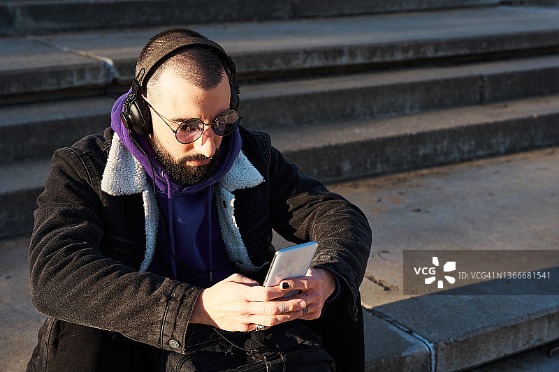 一个戴着耳机的男人坐在户外的台阶上用手机。图片素材
