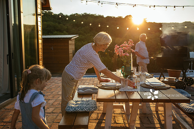 白发苍苍的老女人在户外摆餐桌，他的小孙女在帮她，老男人在他们旁边烧烤图片素材