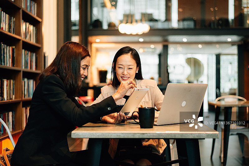 两个亚洲白领妇女在办公室咖啡厅边看笔记本电脑边讨论图片素材