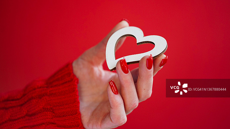 特写的女性手与红色的指甲持有白色木制心脏在红色的背景，复制空间。情人节的概念。美丽的节日卡片。爱。冬天。浪漫。图片素材
