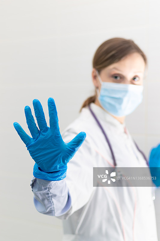 年轻漂亮的女医生戴着医用口罩和手套，拿着听诊器在医院里做手势。有选择性的重点。肖像图片素材