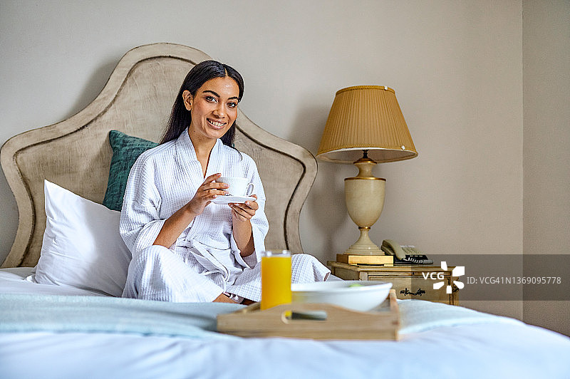 照片中一位迷人的年轻女子盘腿坐在她的床上，享受着她在酒店的早晨咖啡图片素材