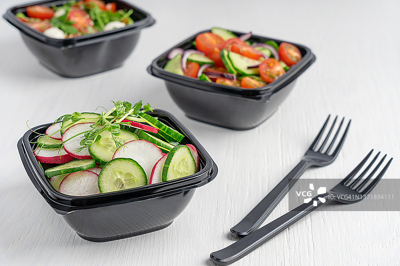 一次性塑料午餐盒，用于食品配送，不同的蔬菜沙拉，服务在白色的木制桌子和叉子图片素材