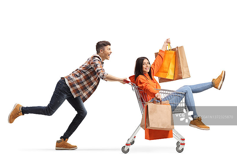 一名年轻男子推着一名年轻女子坐在购物车里，手里拿着购物袋图片素材