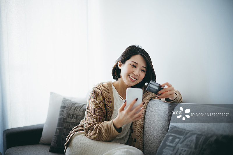 快乐的亚洲年轻女子用她的智能手机和她的信用卡在家里做网上购物图片素材
