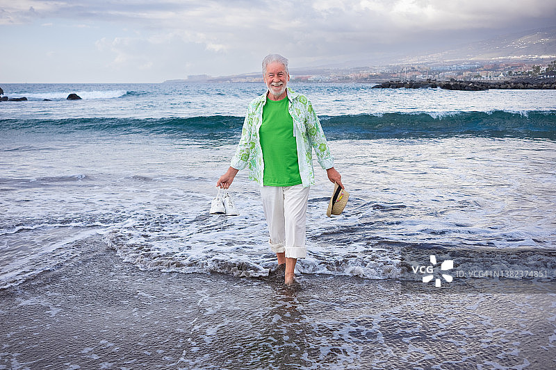 快乐的年老有吸引力的男人赤脚站在海滩的海岸线上，手里拿着鞋子和帽子享受假期的概念轻松和快乐的退休人员图片素材