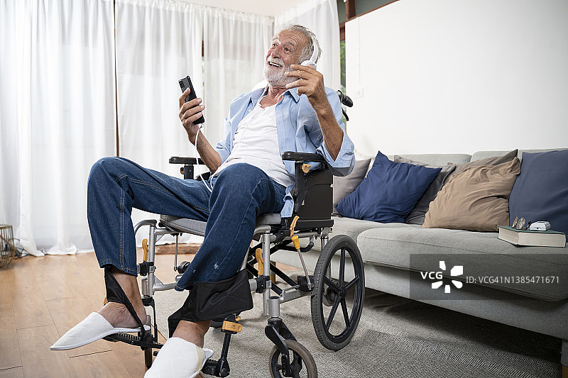 快乐的残疾老人坐在轮椅上用智能手机和耳机在家里的客厅里听他最喜欢的音乐图片素材