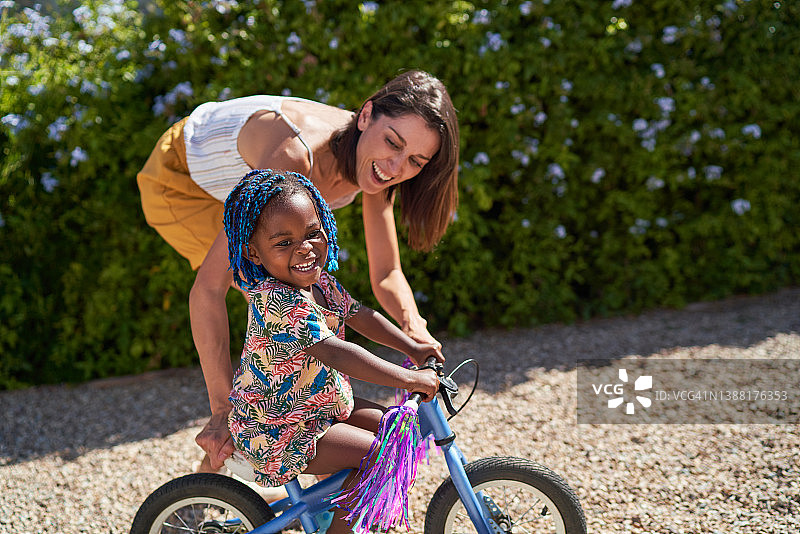 快乐的妈妈在阳光明媚的车道上帮助女儿骑自行车图片素材