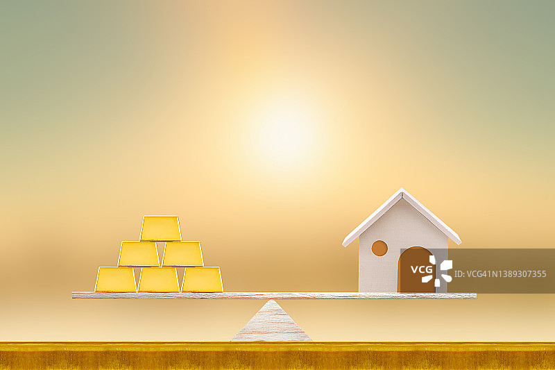 家和堆叠的金条放在磅秤上，平衡放在木头上的阳光背景，储蓄为买新房或房地产和贷款为计划未来的商业投资的概念。图片素材
