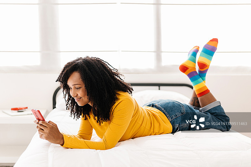 快乐的年轻非洲裔美国妇女使用手机躺在家里的床上。图片素材
