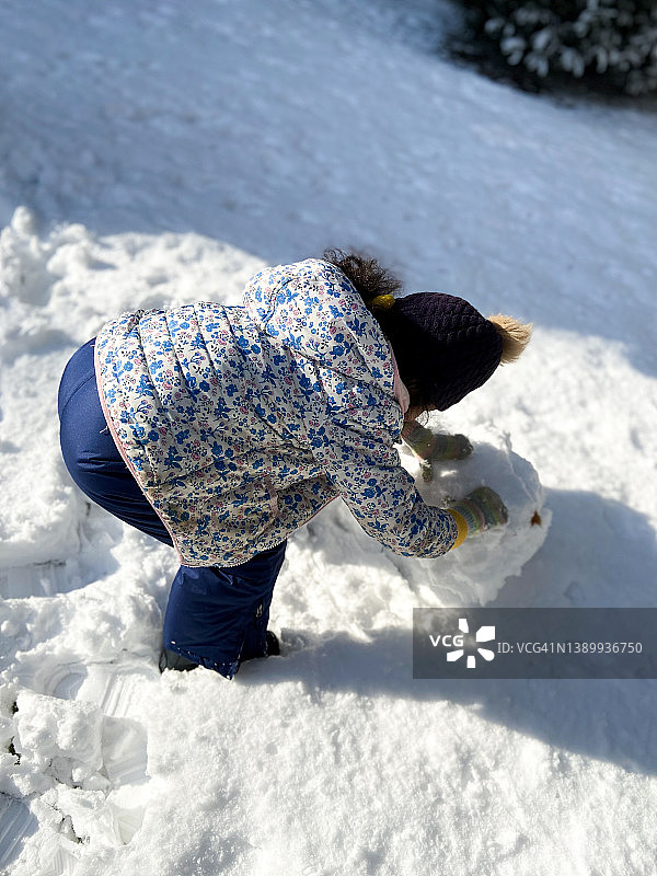 顽皮的混血小女孩戴着花球帽，在一场冬季暴风雪后的新雪中堆雪人图片素材